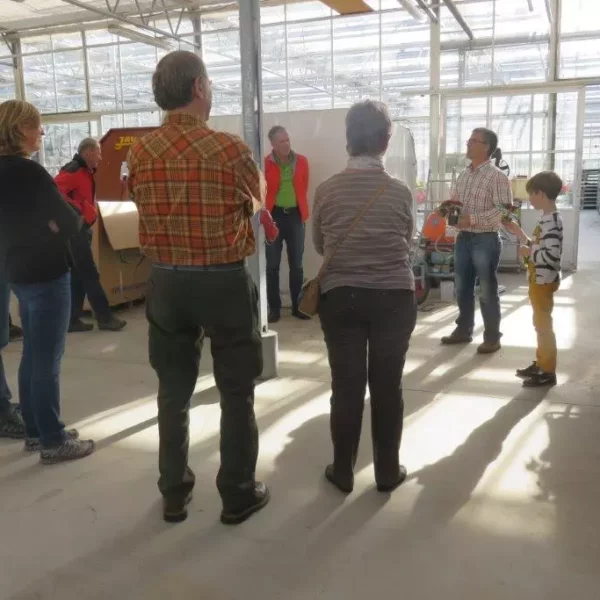 Gruppe von Menschen in einer Produktionsgärtnerei mit Bäumen, Büsche und Pflanzen in Regalen bei einer Führung im Sonnenlicht