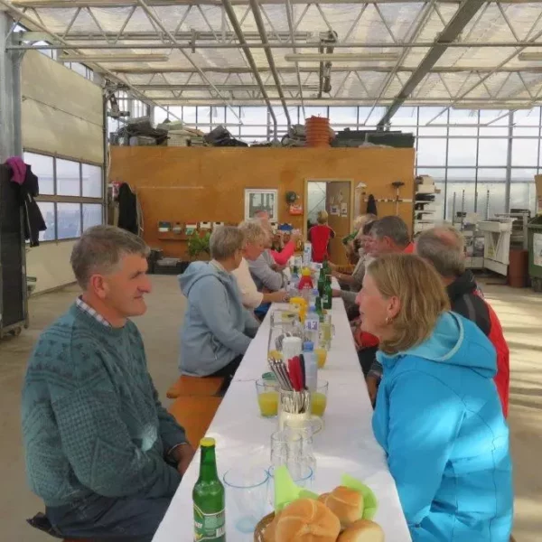 Menschen sitzen an einem langen Tisch mit Getränken und Brötchen in einer hellen Produktionsgärtnerei mit Bäumen und Büsche
