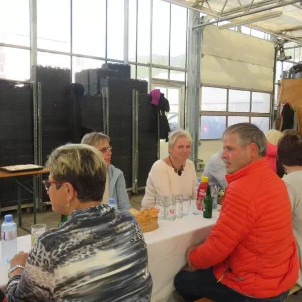 Menschen sitzen an einem langen Tisch in einer Halle mit großen Fenstern, umgeben von Regalen mit Pflanzgefäßen und