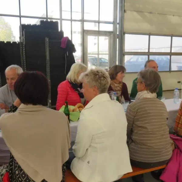 Menschen sitzen an einem langen Tisch in einer Halle, unterhalten sich und trinken Getränke beim Obst- und Gartenbauverein