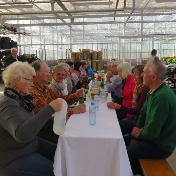 Menschen sitzen an einem langen Tisch mit Getränken in einer hellen Produktionsgärtnerei mit Regalen und Pflanzen im