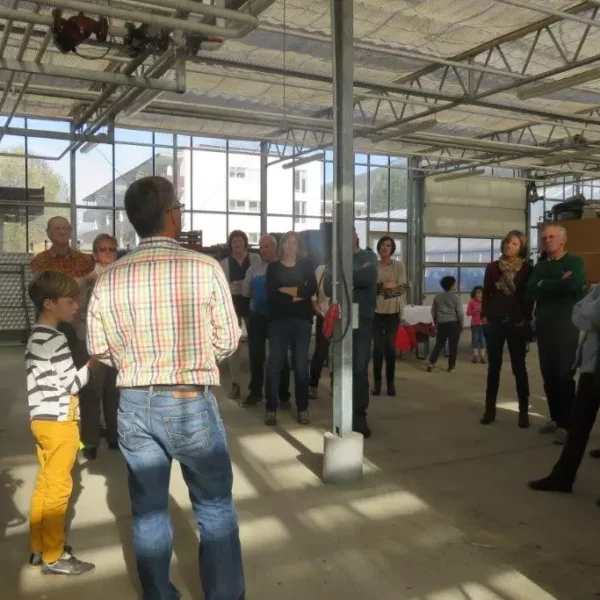 Gruppe von Menschen in einer hellen Halle mit Glaswänden, einige stehen vor einem Mann in kariertem Hemd, der spricht.