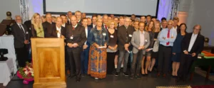 Große Gruppe von Menschen bei einer Veranstaltung, einige halten Urkunden, vor einem Rednerpult mit Blumenarrangement.
