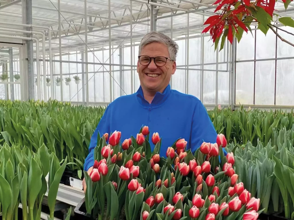Mann in blauer Jacke lächelt in Gewächshaus mit roten Tulpen und grünen Pflanzen im Hintergrund in Sankt Johann Pongau